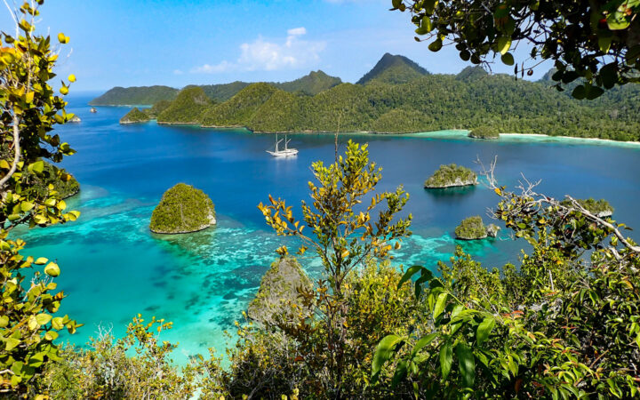 looking down at a beautiful desolate indonesia cove with a small ship anchored in the middle of it and jungle surrounding it.