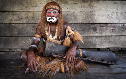 West Papuan tribesman sits with a wooden drum in front of a weathered wood-plank wall during the Spice Islands & Raja Ampat small ship cruise in Indonesia.