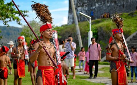Men in red loincloths and red headbands with tall brown feathers march in order beside a stone fort on a sunny day during the Spice Islands & Raja Ampat small ship cruise in Indonesia.