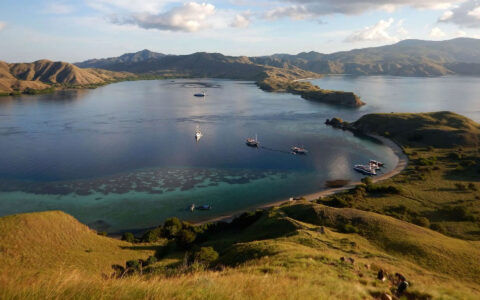 Aerial view of Indonesian archipelago with blue & turquoise sea, hills draped in green & gold and small ships sitting at anchor.