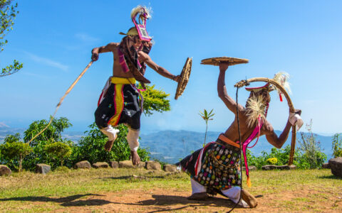 2 men in colorful native garb on a mountaintop do a demonstration fight dance with wood stalks & shields in the Indonesian sun.