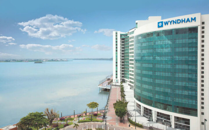 Exterior of Wyndham Guayaquil Hotel, multiple-story glass-encased building overlooking a large scenic river and a riverside walkway on a sunny day.