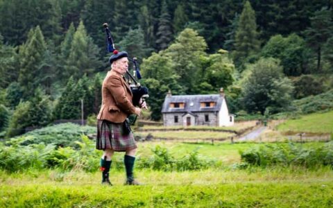 Kilted Scottish bag piper plays his instrument among bright green grass with forest & a small country home in the background.