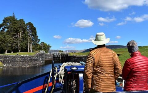 Man & woman in bright down jackets stand at the front of a hotel barge as it approaches a loch on a sunny day during the Classic Scotland Barge Cruise.