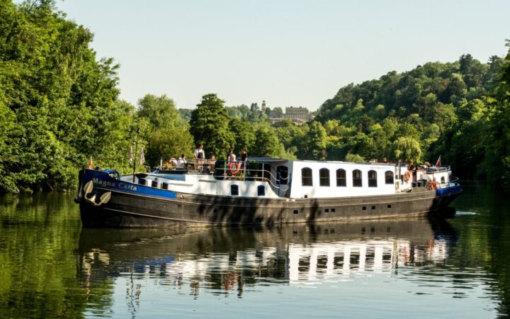 Black, white & blue-painted Magna Carta barge cruises the River Thames in calm waters with tree-lined riverbanks.