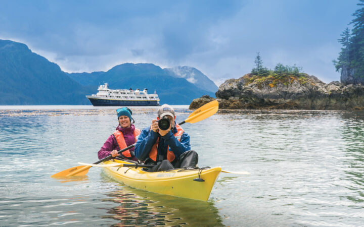 An Alaska small ship cruises while in a double yellow kayak a female paddles while her male partner snaps a photo.