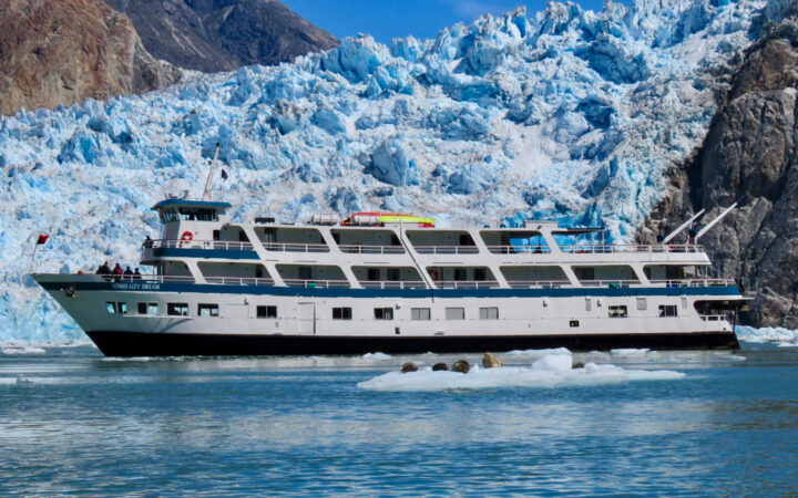 Admiralty Dream cruise ship with white decks sits parked in front of a large blue glacier on a sunny day in Alaska.