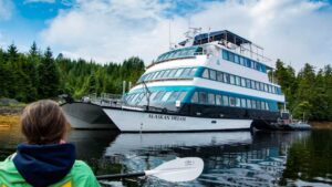 Kayaker paddling calm sea back to the 4-deck, blue & teal catamaran small ship Alaskan Dream on anchor by green forest.