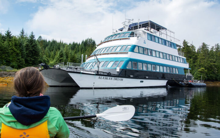 Kayaker paddling calm sea back to the 4-deck, blue & teal catamaran small ship Alaskan Dream on anchor by green forest.