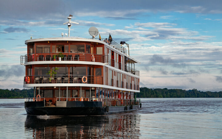 Small river cruise ship tan and wood in design with white roofs and a black bottom cruises the amazon on a calm day