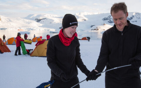 Man & woman in black clothing put together tent poles with others setting up tents in the snow behind them, in Antarctica.