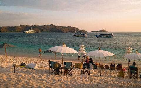 White-sand beach set up with ornate umbrellas & camping chairs by turquoise water with 3 small ships offshore at sunset.