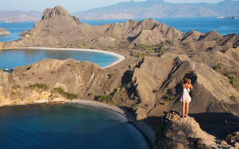 A female guest from a Komodo national park cruise stands on top of the jagged volcanic mountains to take a photo of the dramatic landscape.