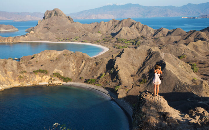 A female guest from a Komodo national park cruise stands on top of the jagged volcanic mountains to take a photo of the dramatic landscape.