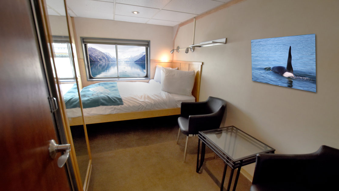 Category A cabin on Baranof Dream Alaska small ship with double bed in white linens, picture window, brown chairs & glass coffee table.