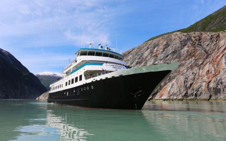 Chichagof Dream ship bow seen from water level with black hull, white upper decks & aqua trim on a sunny day in Alaska.