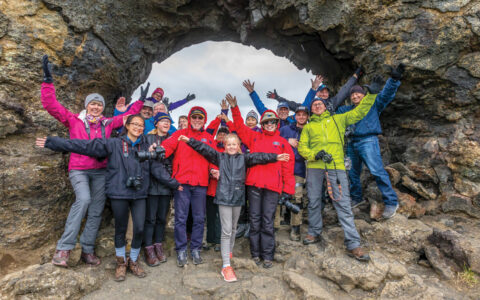 Group of Iceland Circumnavigation cruise guests stands with arms outstretched for a photo opp under a gray lava arch.