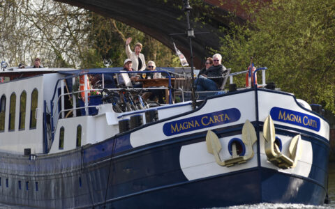 Classic England River Thames barge cruise guests wave from the bow deck as their small blue & white ship cruises under a bridge.