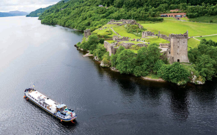 Aerial view of Urquhart Castle set along the Great Glen waterway, surrounded by bright green trees and grass with a hotel barge ship cruising by.
