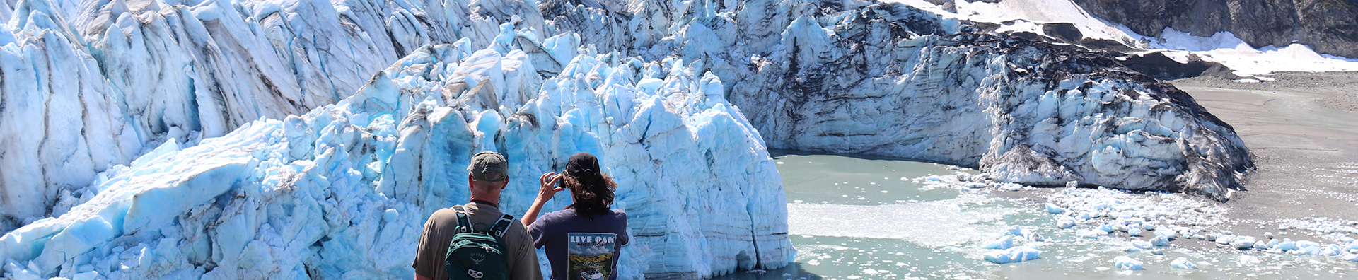 Two travelers in t-shirts and hats face a massive blue and white glacier with the female taking a photo of it