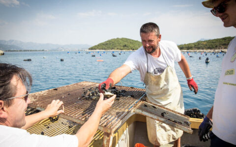 A man in an apron passes mussels to another man in sunglasses with the ocean behind them
