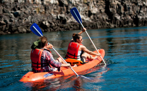 Tandem kayaking pair paddles an orange boat beside rocky cliffs in calm water on an Evolution Galapagos cruise.