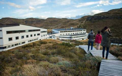 Explora Connects travelers on a boardwalk by rectangular white hotel among pampas grassland in Torres del Paine National Park.
