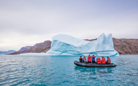 Black inflatable Zodiac dinghy with Arctic travelers motors past a large iceberg in calm water on an Iceland to Greenland cruise,