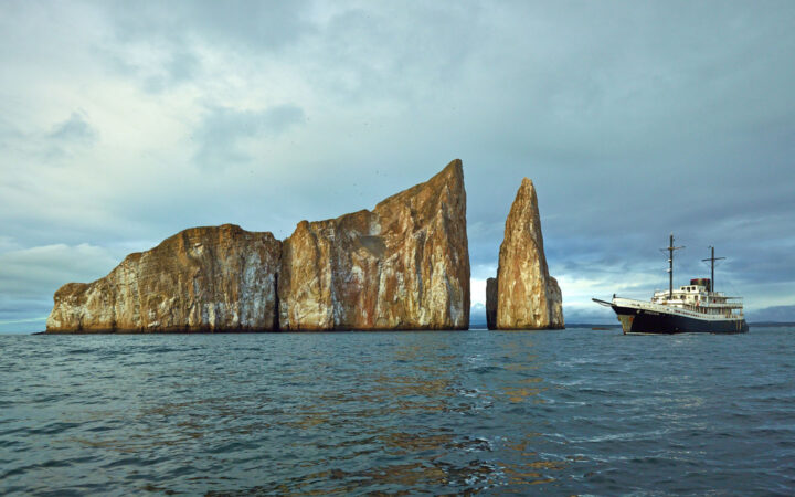 White & blue Galapagos small ship Evolution floats beside Kicker Rock, a tan volcanic cone that reaches up from the ocean.