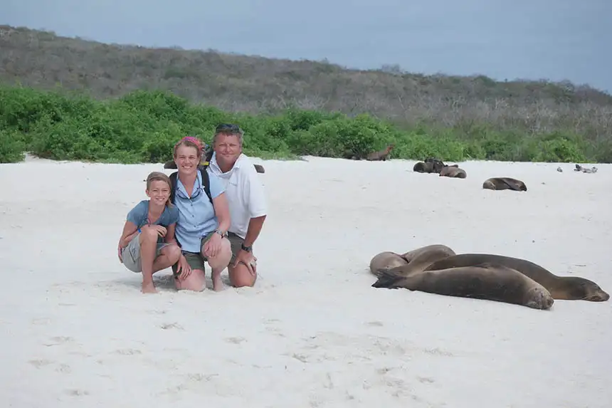 A family of three squats on a white sand beach next to Galapagos sea lions resting