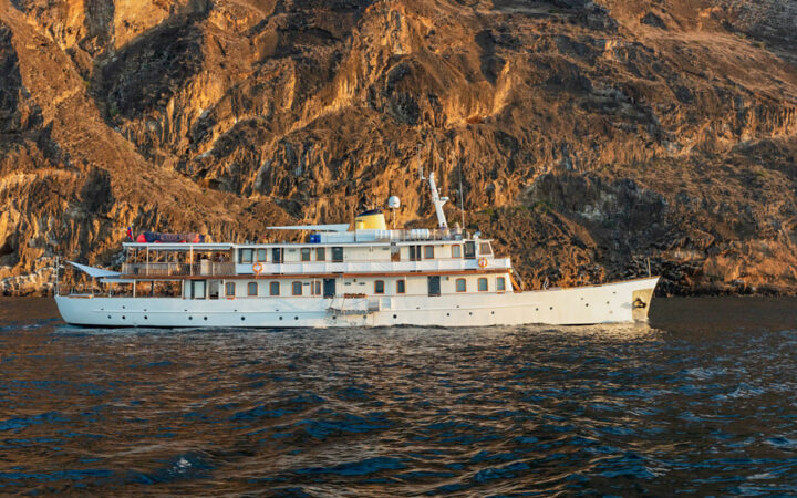 Grace boat in Galapagos with white hull & 2 upper decks, wood finishes & many windows, sits beside golden cliffs in calm sea.