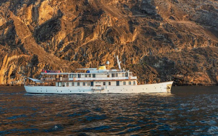 Grace boat in Galapagos with white hull & 2 upper decks, wood finishes & many windows, sits beside golden cliffs in calm sea.