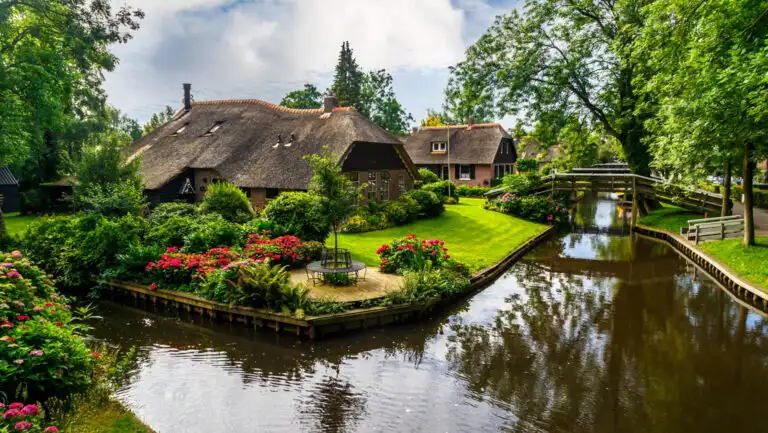 Glassy canal flows beside bright green grass, gardens of red flowers & shake-wood roofs on a sunny day of a Holland river cruise.