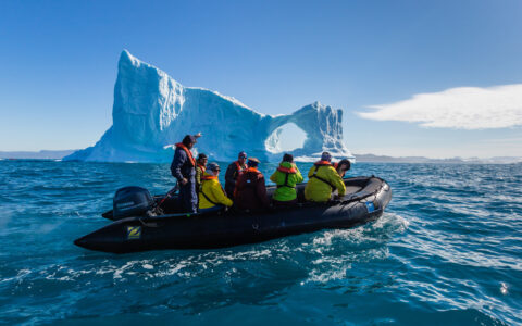 Black Zodiac boat with Greenland travelers in colorful jackets cruises past a large iceberg with a circle cut out.