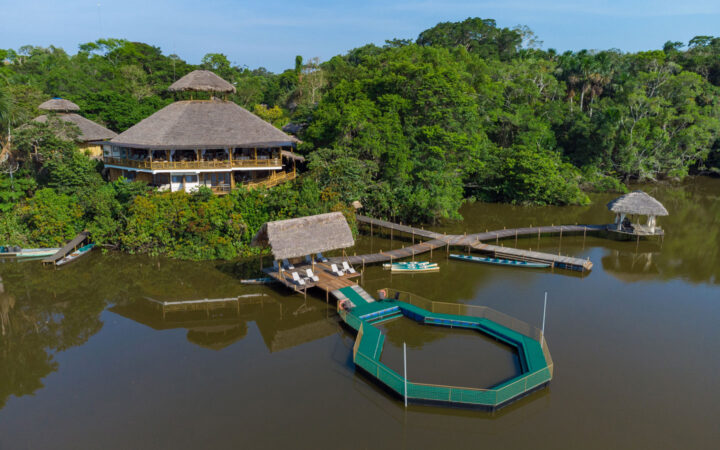 Aerial view of La Selva Lodge with hexagonal thatch-roof buildings beside a lake with 4-way wood dock & green-circled natural pool.