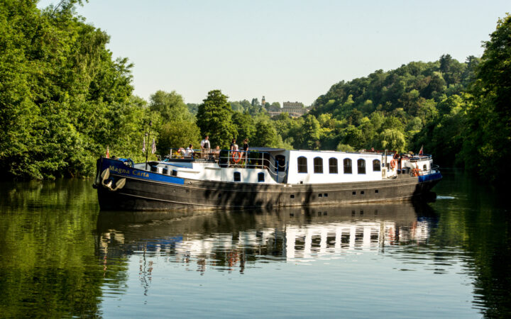 Black, white & blue-painted Magna Carta barge cruises the River Thames in calm waters with tree-lined riverbanks.