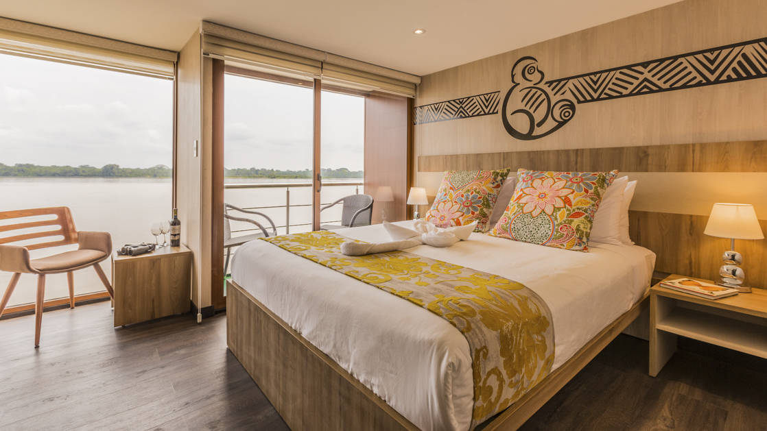 Standard Cabin aboard Manatee Amazon Explorer with floor to ceiling windows, sliding glass door out to private balcony with nightstand and reading light.