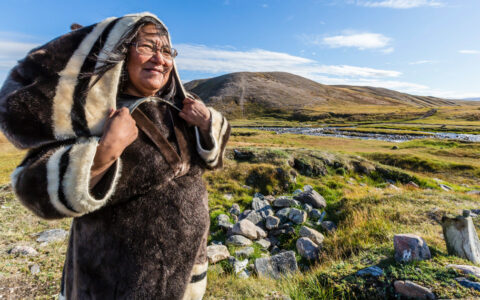 Native woman in hooded overcoat made from dark animal fur stands in green tundra backed by mountains on a sunny day.