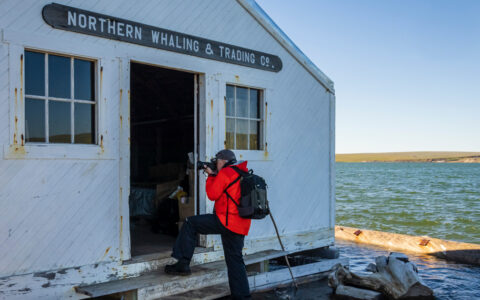 Northwest Passage traveler in red jacket stands outside door of historic whaling company building at water's edge & takes photos.