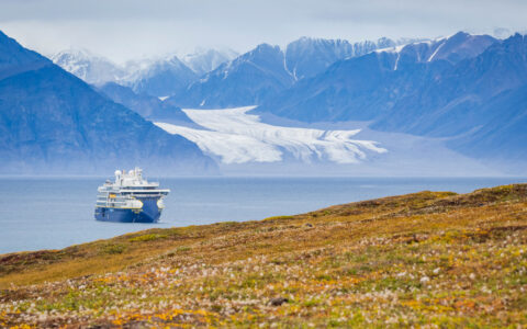 Small, modern, blue & white expedition ship sits in calm fjord beside rocky mountains, white glacier & green riverbanks.