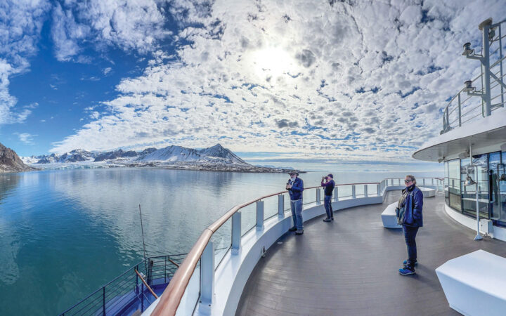 3 travelers stand on deck of a modern ship viewing rocky & snowy peaks & a large white glacier on a Norwegian Discovery cruise.