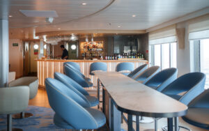Long table with tall blue swivel chairs leads to modern, lit-up bar by wall of windows on National Geographic Explorer ship.