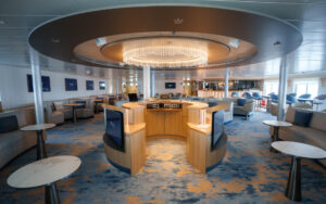 Interior lounge on National Geographic Explorer ship with round dais under chandelier, surrounded by padded benches & small tables.