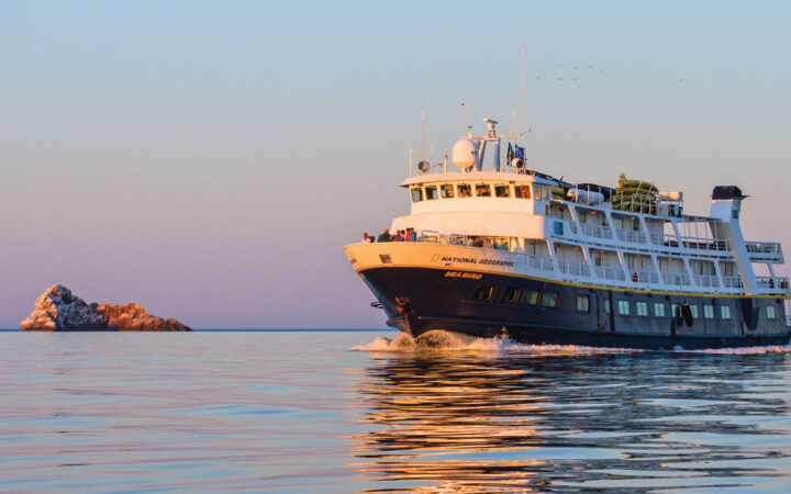 National Geographic Sea Bird small ship with dark blue hull & white upper decks cruises through calm seas at sunset.