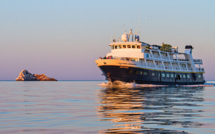 National Geographic Sea Bird small ship with dark blue hull & white upper decks cruises through calm seas at sunset.
