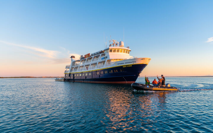 National Geographic Sea Lion small ship with dark blue hull & white upper decks sits on anchor as Zodiac cruises by at dusk.