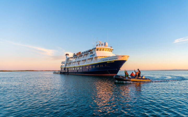 National Geographic Sea Lion small ship with dark blue hull & white upper decks sits on anchor as Zodiac cruises by at dusk.