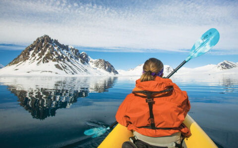 Girl in orange jacket paddles a sit-on-top yellow kayak in glassy water beside snowy mountains on a Norwegian Discovery cruise.