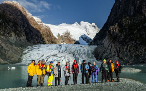 Chilean Patagonia cruise travelers in bright coats stand on stony shore in front of an icy blue glacier & snowy peaks behind.