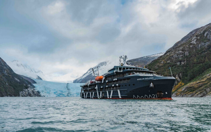 Magellan Explorer dark blue small ship sits in calm water by blue glacier on cloudy day in Patagonia.